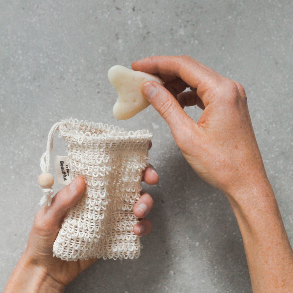 Hands holding an open sisal soap saver pouch over a stone surface, with a small bar of soap being lifted out