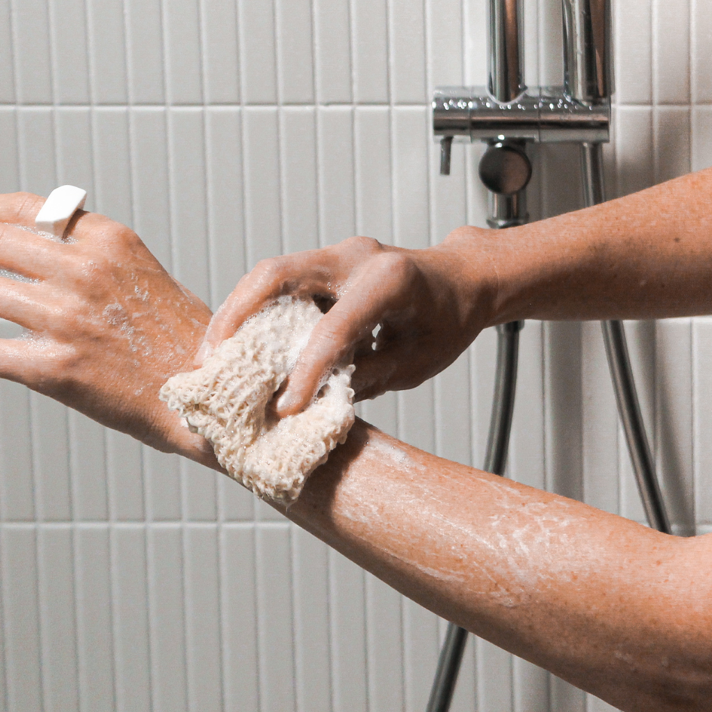 Person scrubbing a wet forearm with a soapy sisal pouch in a tiled shower, creating lather